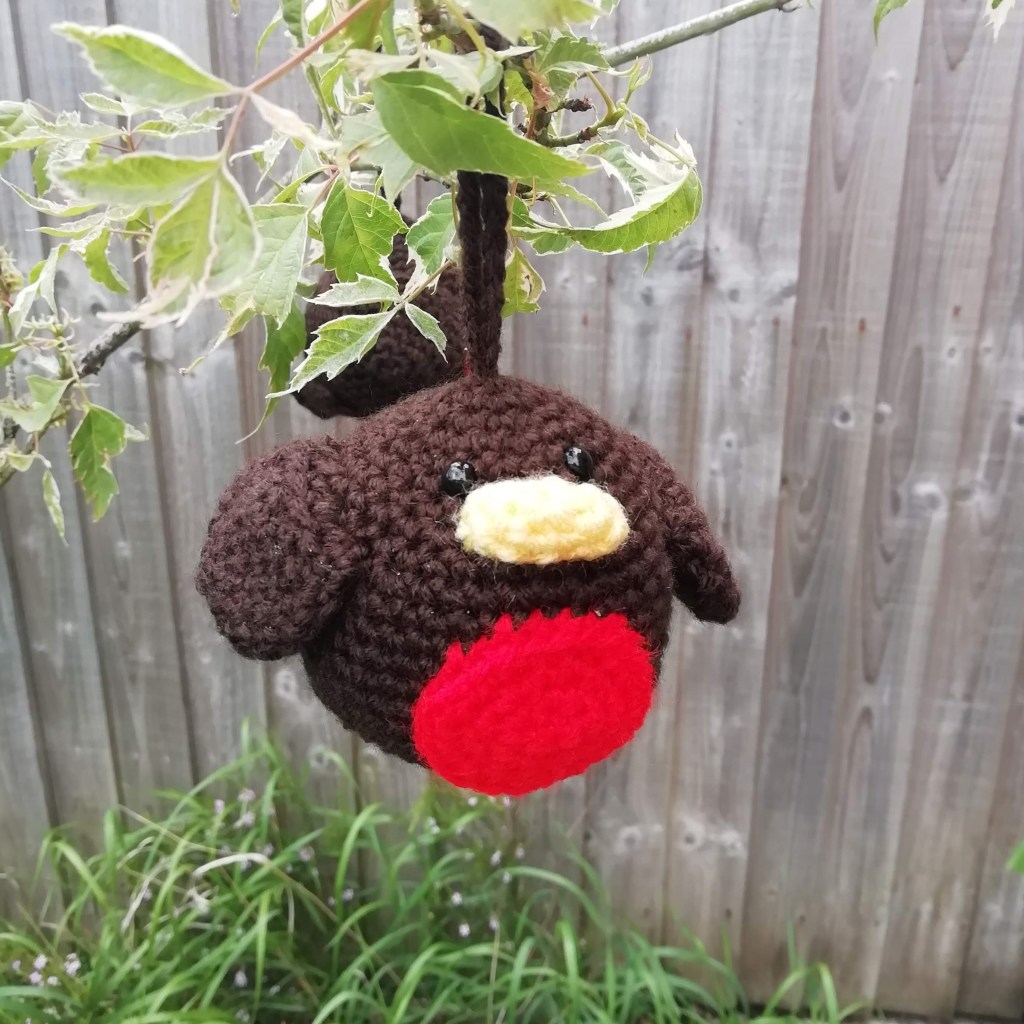 A round crochet robin. It is brown with a circular red breast and yellow beak. It is hanging on a tree with a brown crochet loop.